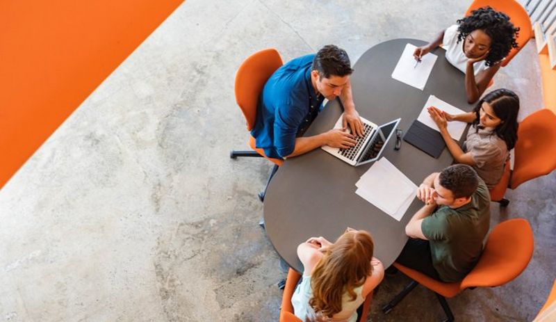 Overhead shot of office workers sat round an oval-shaped table on orange chairs, spread with papers and a laptop computer.